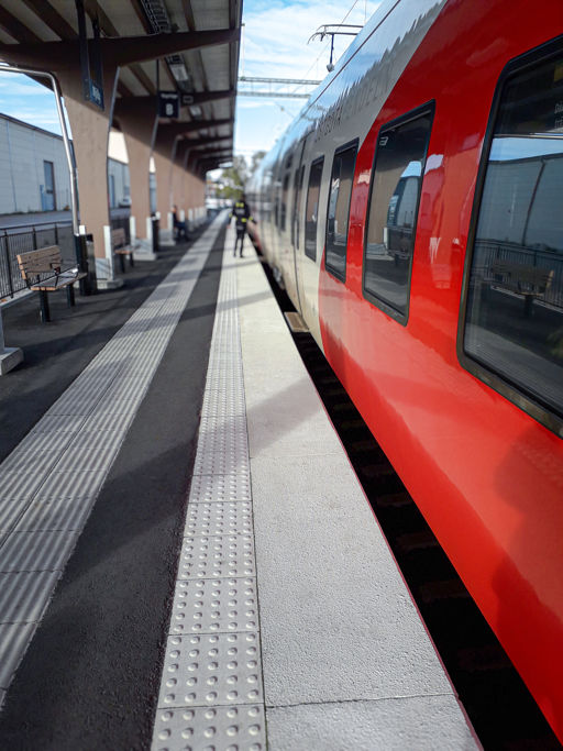 Vy över en perrong med ett tåg längs perrongen med vita kantplattor kantade av ledstårk av vita superplattor med både kupol- sinusmönster.
