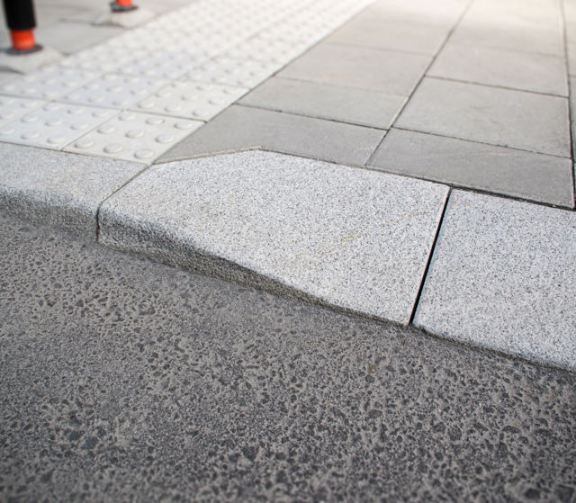 Portstens i natursten;kompletterad med taktila betongplattor med orienteringsmönster samt släta betongplattor i gångyta – i övergång mellan gångbana och körbana.